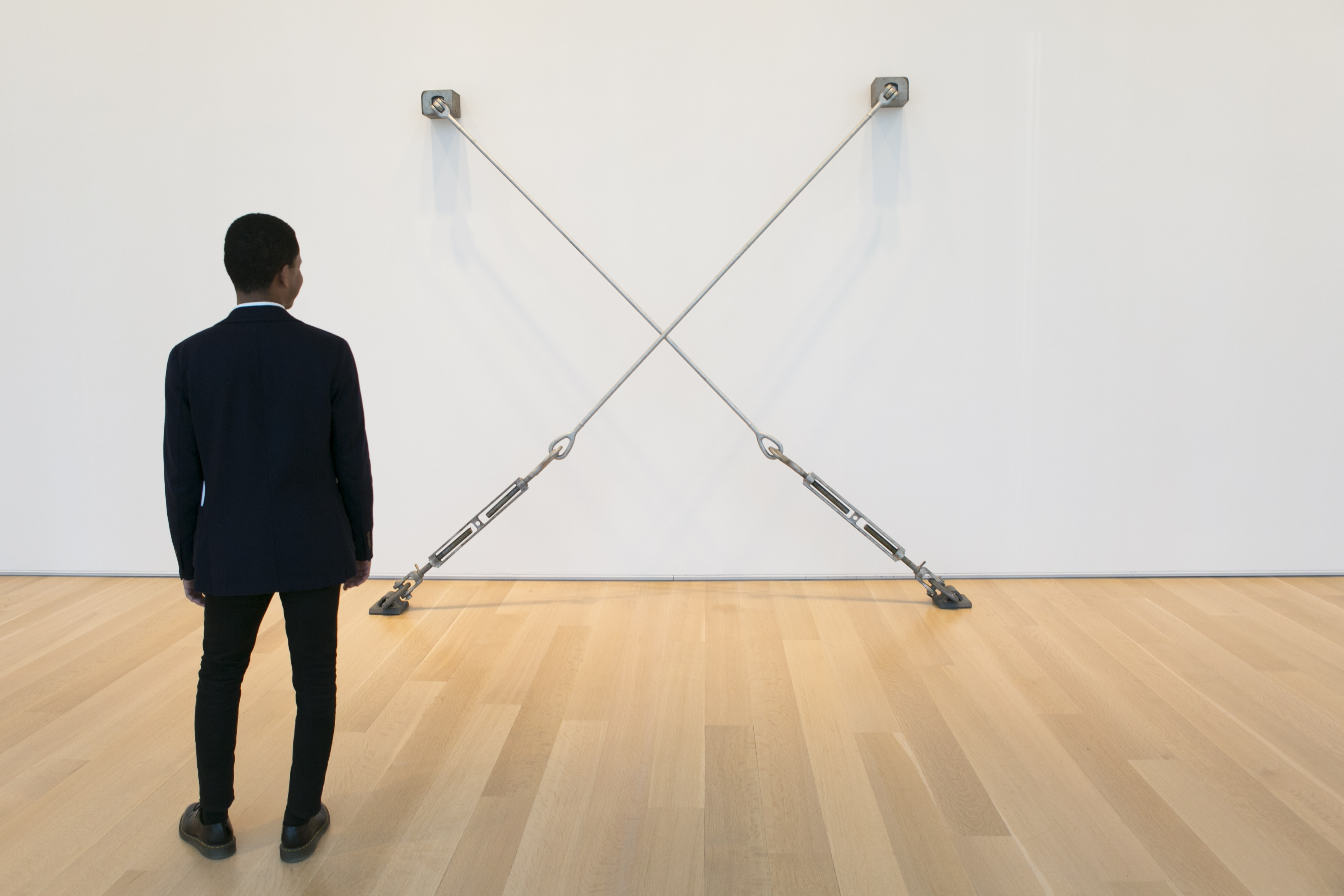 A person views an artwork that uses metal container lashing bars hung installed on a white wall and a wooden floor.
