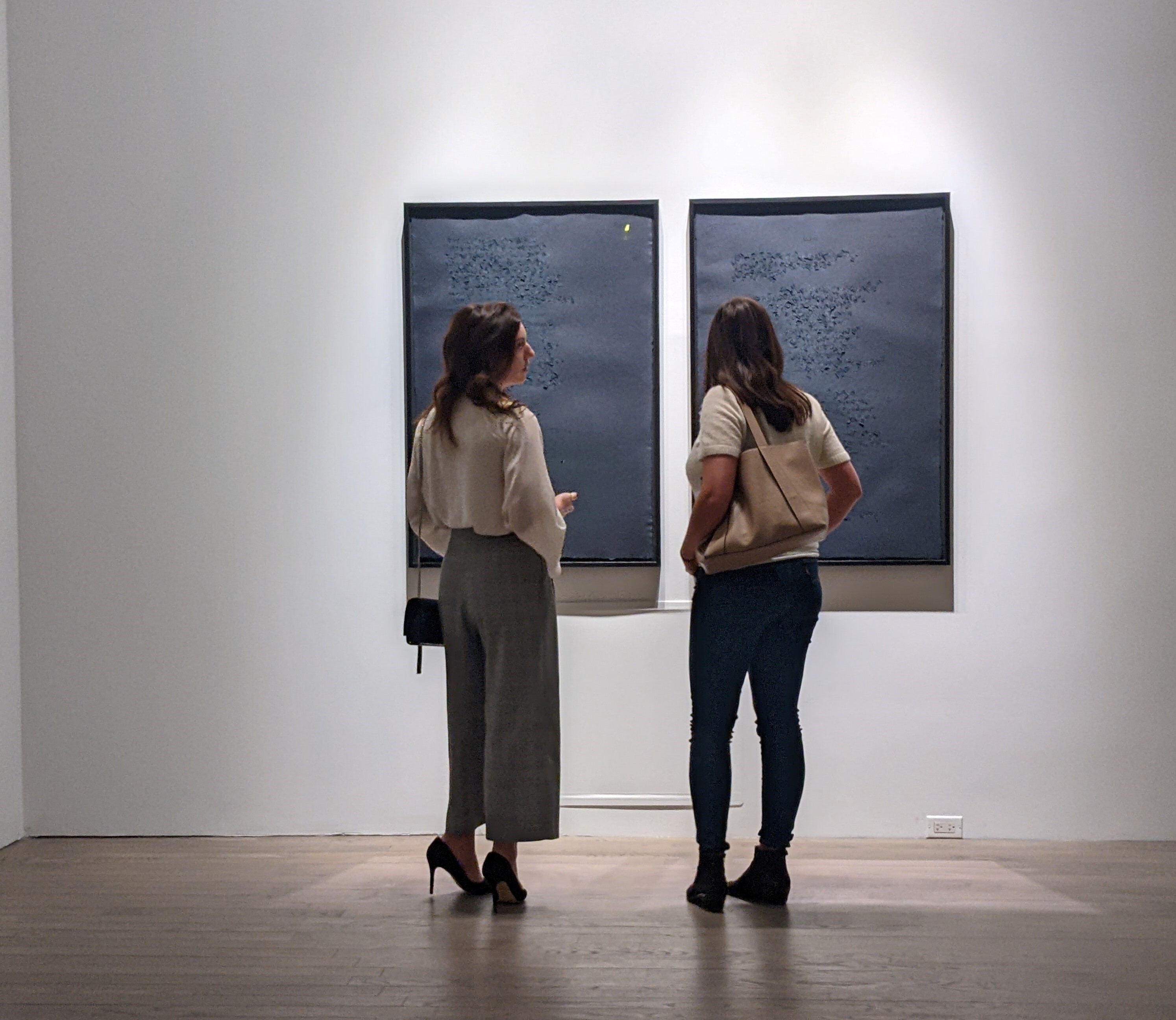 Two women view a diptych artwork on a white gallery wall.